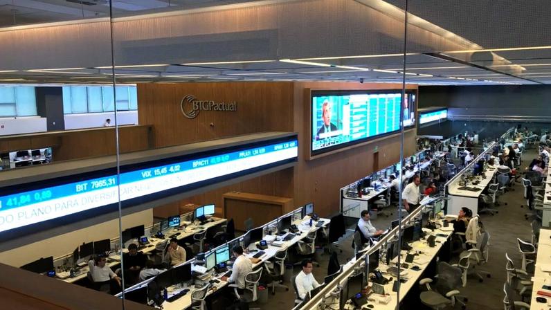 Corretora de valores BTG Pactual, com funcionários em mesas e grandes telas exibindo dados financeiros.