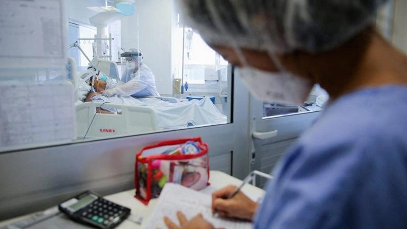 Profissionais de saúde em hospital: um escreve e outro assiste paciente em leito, separados por vidro.