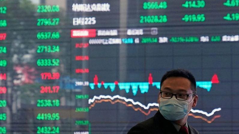 Um homem de máscara está em frente a um grande painel digital exibindo gráficos e números do mercado financeiro.