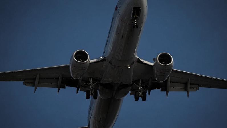 Avião comercial prateado, visto de baixo, com trem de pouso estendido, decolando contra um céu azul claro.