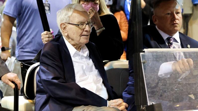 Warren Buffett em carrinho de golfe, com motorista e mulher de máscara ao fundo.