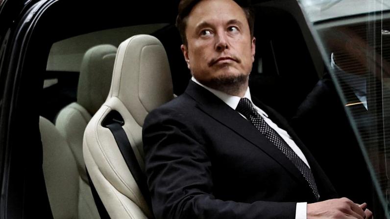 Elon Musk sentado no banco traseiro de um carro, de terno, olhando pensativo para a direita.