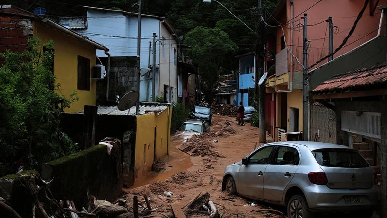 Rua cheia de lama que chega a cobrir alguns carros estacionados e ao fundo árvores