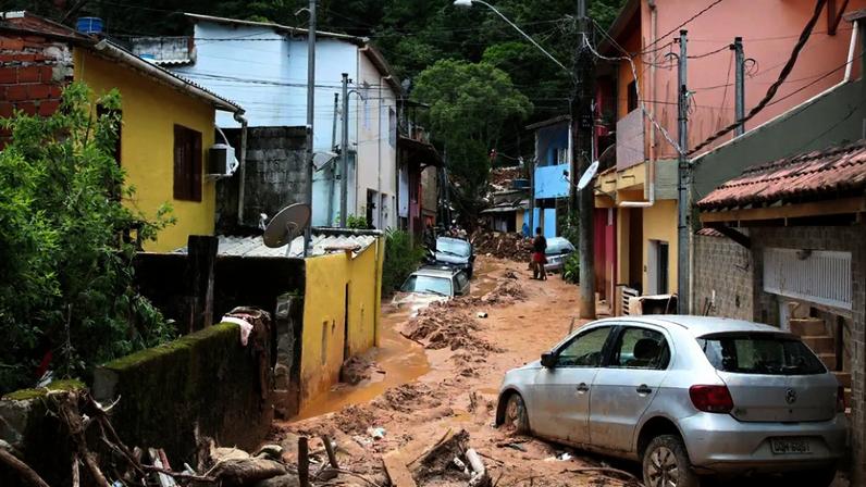 Rua cheia de lama que chega a cobrir alguns carros estacionados e ao fundo árvores