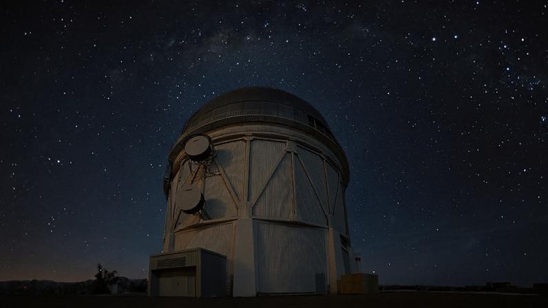 Observatório astronômico com paredes metálicas brancas e cúpula cinza, situada ao centro de uma paisagem com céu estrelado