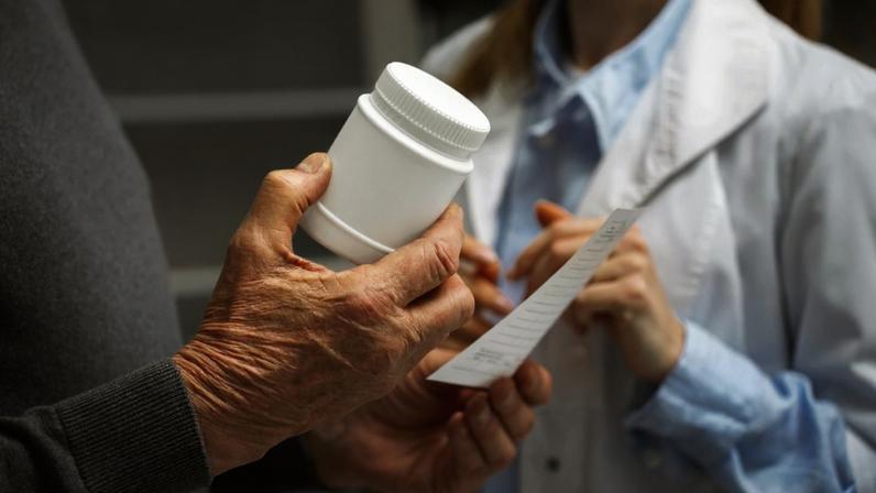 Foco nas mãos de duas pessoas, uma segurando um pote de medicamentos e um papel com a receita médica e outra ao lado de jaleco branco gesticulando