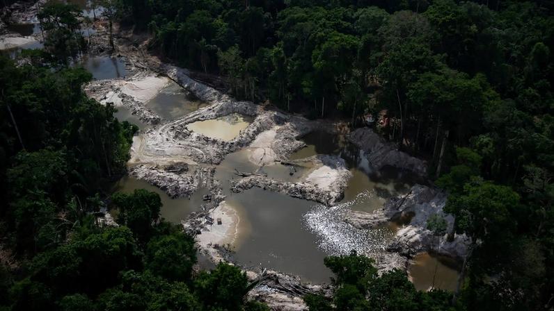 Região de mata afetada pelas práticas ilegais de mineração, mostra uma abertura no meio das árvores da floresta que formou um vale de lama acinzentada e grandes poças de água.