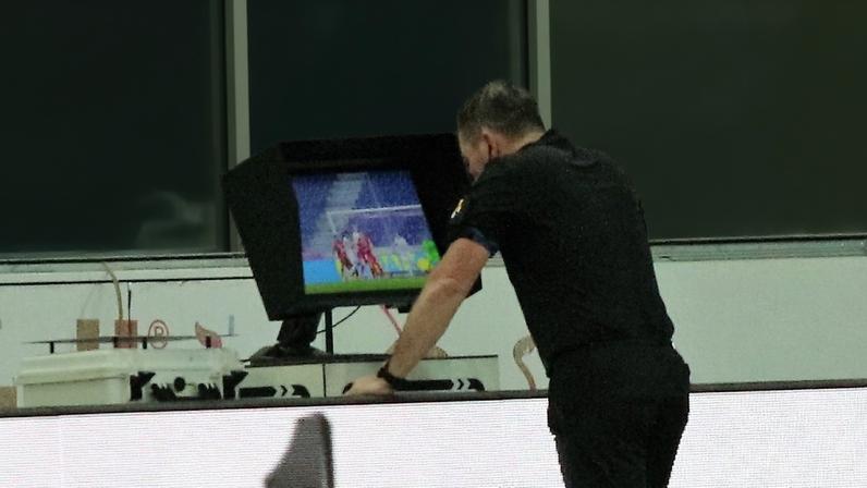 Árbitro de futebol com uniforme preto, visto de costas, consulta o monitor de vídeo do VAR que mostra um lance da partida, enquanto é observado por torcedores