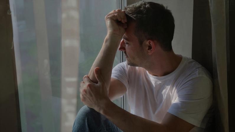 Um homem adulto em um retrato de maio corpo, posicionado de lado, pensativo. A luz entra por uma janela próxima e projeta sombras, como se fossem grades ou persianas, sobre o rosto e o corpo do homem. As sombras atravessam o rosto dele. Parte do rosto fica iluminada, enquanto a outra permanece sombreada.