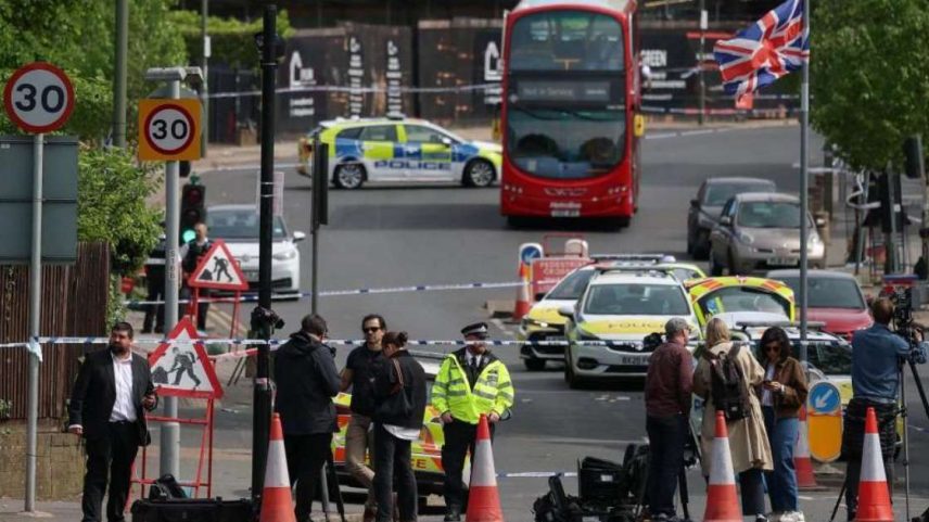 Uma cena de rua mostra membros da imprensa trabalhando perto de um cordão policial, em frente a carros da polícia e um ônibus estacionado, enquanto uma bandeira britânica tremula acima deles