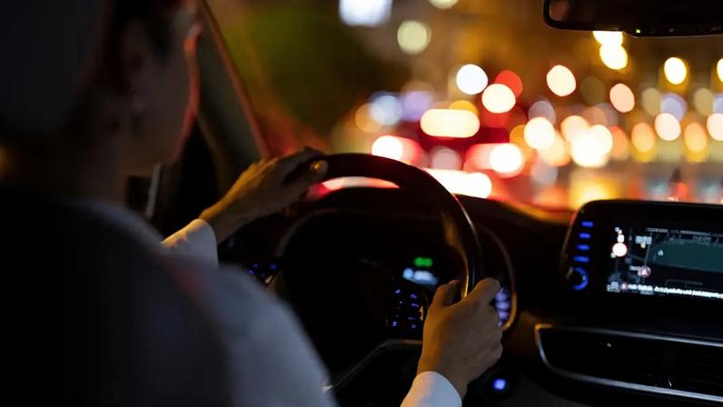 Fotografia de uma mulher dirigindo um carro à noite com luzes coloridas da cidade e tela de navegação iluminando o interior.