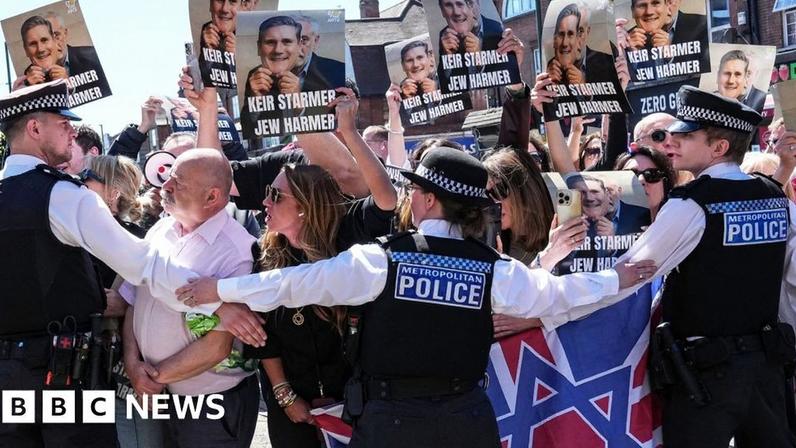 Protesters holding up placards with Keir Starmer's face printed on them being held back by police in Golders Green today 3oth April 2026