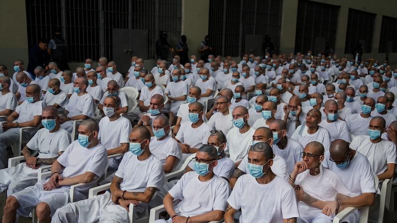 A large group os men sitting on rows of chairs wearing prison uniforms and face masks with their wrists and ankles chained
