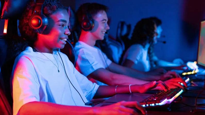 Grupo de adolescentes multirraciais em fones de ouvido jogando videogames em um clube de videogames com iluminação azul e vermelha, teclado e mouse com iluminação