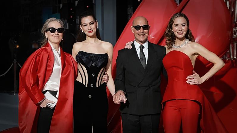 Meryl Streep, Anne Hathaway, Stanley Tucci and Emily Blunt pose on the red carpet in London. Streep is wearing sunglasses, a long, red, satin jacket, a cream blouse and black trousers. Hathaway has her hair up and is wearing a black embellished corset. Tucci is wearing a pinstriped black suit, white shirt and tie, along with sunglasses. Blunt has a red two-piece on an a jewelled necklace.