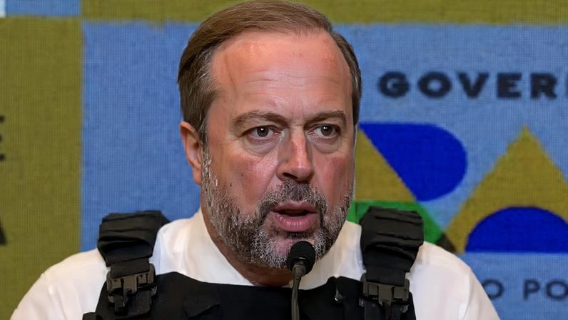 Homem fala em coletiva de imprensa. Veste camisa branca e colete tático preto com crachá escrito “Dr. Silveira Minas e Energia”. Atrás, painel com logotipo “Governo do Brasil” nas cores da bandeira.