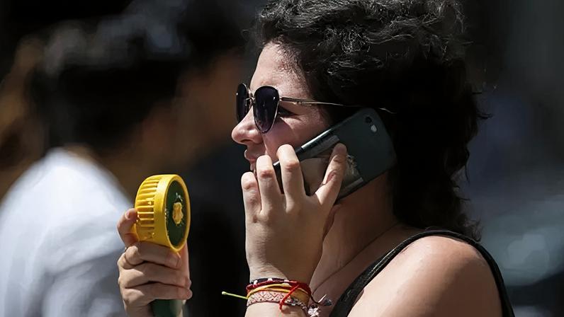 Getty Images A young woman wearing sunglasses talks on the phone while using a portable fan to keep herself cool.