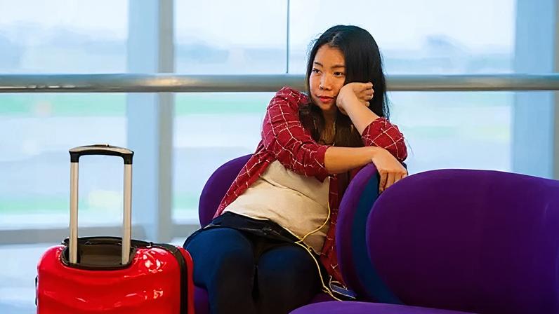Getty Images Young woman in red checked shirt rests her chin on her hand. She is sitting in an airport waiting room with a red case next to her