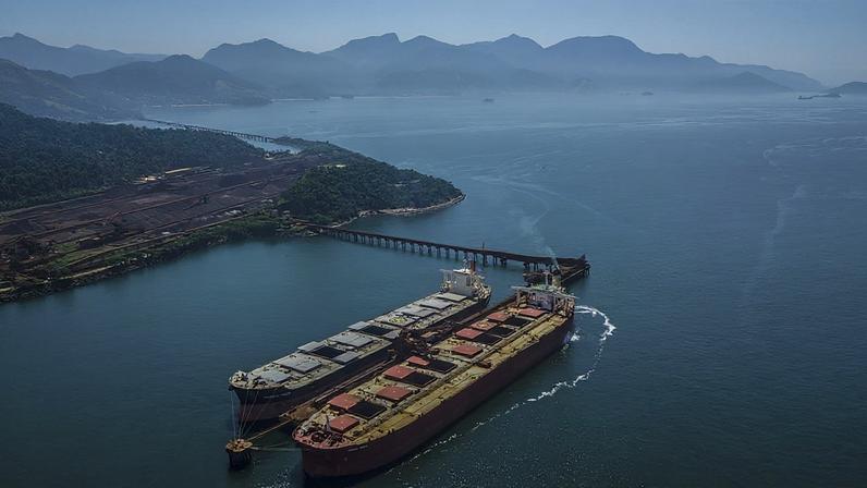 Navios graneleiros em porto industrial, com pátio de minério, montanhas e mar.