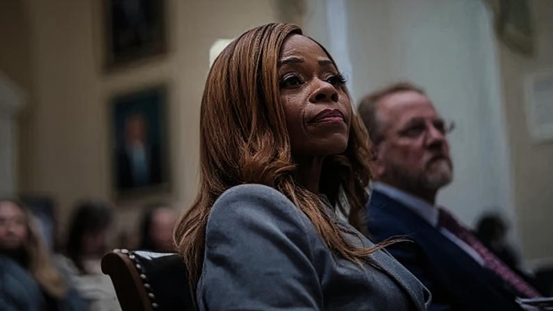 Getty Images A woman with reddish-brown hair looks ahead in a grey suit. She is sitting in a brown chair. In the background, other people sitting in chairs can be seen, but are blurry.