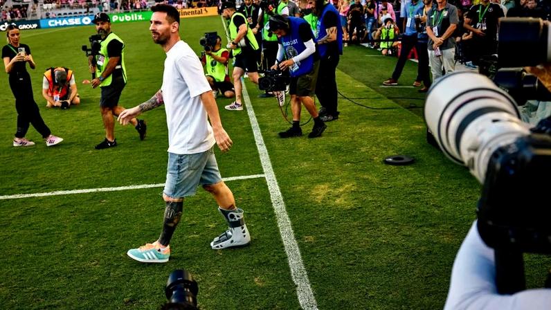 Lionel Messi, de branco, caminha por um campo de futebol, cercado por fotógrafos e outras pessoas.