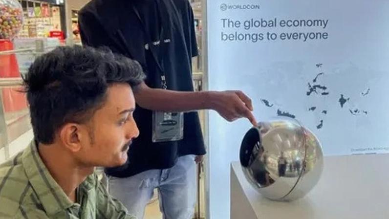 Reuters A man in India sitting before an earlier World device, a silver orb, to have his eye scanned as a form of human identification. He has dark hair and a green shirt, and looks straight into the orb, on a desk in front of him, while another man points to it.