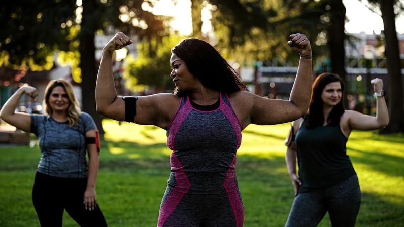 Foto colorida de três mulheres fazendo sinal de força com os braços, vestidas com trajes de exercício físico - Metrópoles.