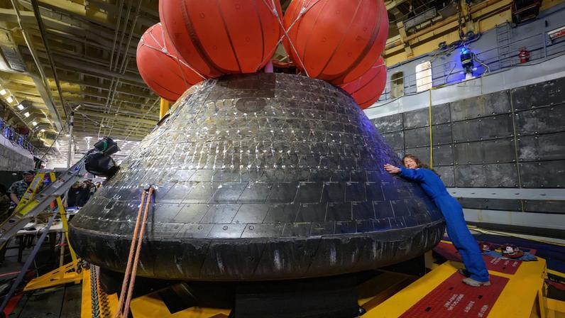 NASA astronaut Christina Koch hugs the Orion spacecraft with her face toward the camera. She is wearing a bright blue jumpsuit. Orion is a rounded cone shaped spacecraft. Its exterior is a sooty black. There are several large orange balloons attached to its top. Orion looks about twice Koch's height.