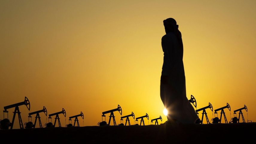 Uma foto em preto e branco mostra a construção de uma torre rudimentar de extração de petróleo em uma região desértica, com cerca de dez pessoas ao redor