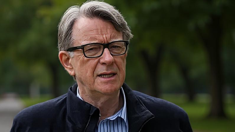 Lord Mandelson has short grey hair swept to one side and is wearing black-rimmed glasses. He has a dark blue jacket on and white and blue striped shirt underneath