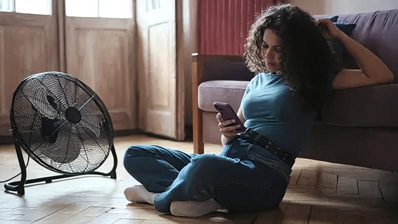 Getty Images A young woman scrolls on her phone as she sits on the wooden floor of her house in front of a free standing black fan.