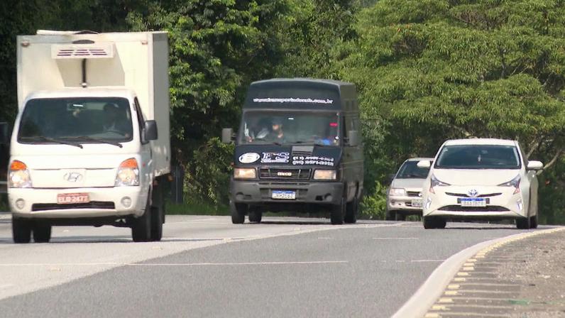 Rodovia de pista simples com tráfego moderado, mostrando um caminhão, uma van e um carro seguindo pela estrada cercada por árvores