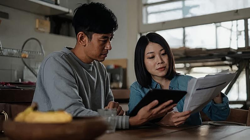 Getty Images A young couple sit in their kitchen looking at bills. The woman is holding papers and the man is holding a tablet device