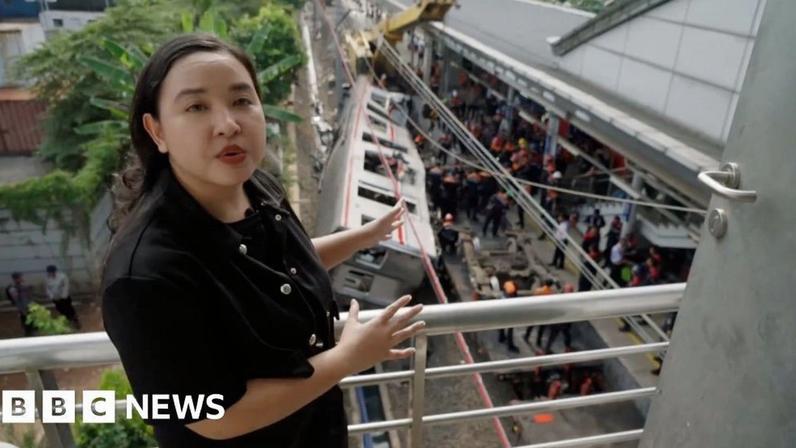 Liza stands above the train crash site and points to it as she speaks