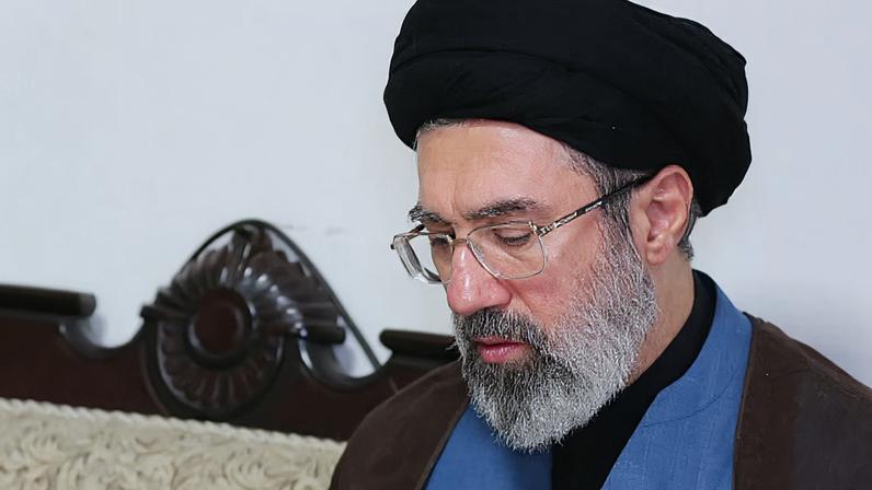 A imagem mostra Mojtaba Khamenei sentado em um ambiente interno, vestindo um manto escuro sobre uma roupa azul e um turbante preto. O fundo exibe parte de uma namoradeira de madeira esculpida, com detalhes curvos e ornamentados, atrás de um estofado claro.