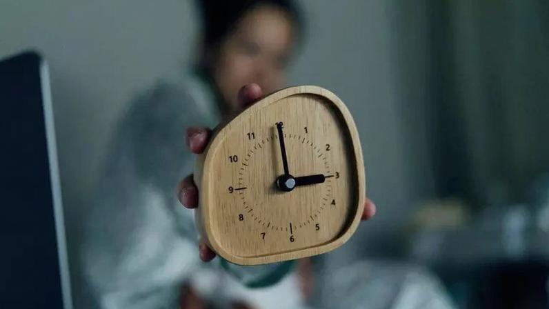 Pessoa sentada na cama mostra um despertador marcando três horas