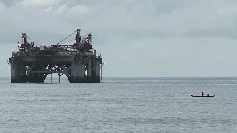 Plataforma de petróleo no mar ao fundo, com uma pequena embarcação com uma pessoa remando em primeiro plano, sob um céu nublado