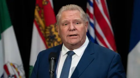 Getty Images Doug Ford stands before a black microphone. He is wearing a navy suit, white shirt and blue tie. He is looking straight ahead. Behind him are several colourful flags.