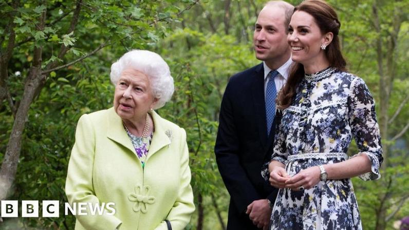 Queen Elizabeth II, wearing a lime green coat, looks off to the side of the camera, as William and Catherine, both smiling, follow her gaze. They are standing in a garden.