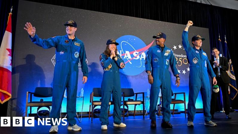 This indoor photograph shows the four Artemis II crew members standing on a stage in matching royal-blue flight suits. Behind them is a large dark backdrop with a prominent NASA logo and starry design, with national flags placed at the sides. One astronaut waves, another appears to speak into a microphone, and another raises a fist in celebration. The lighting is bright and theatrical, and the mood is triumphant, as if the crew are being welcomed back after their mission.