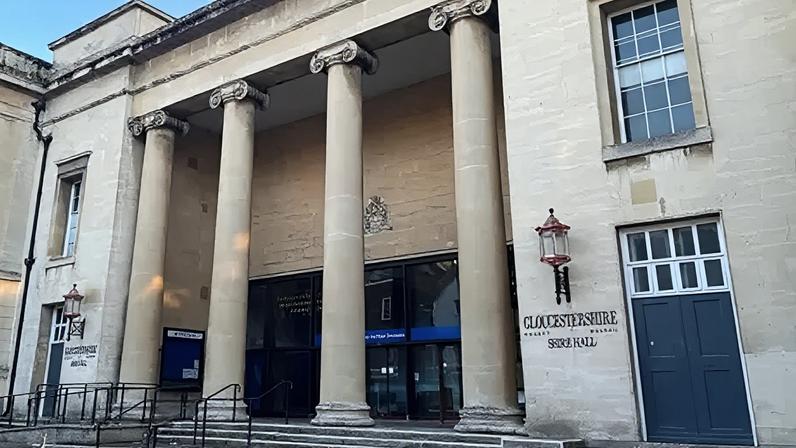 Carmelo Garcia The frontage of Gloucestershire County Council's Shire Hall. The front of the building has four floor to ceiling concrete pillars at the top of the steps, with the entrance doors behind them. The name of the building is written on both sides of the pillars. The sky is visible at the top of the photo, which is bright blue.