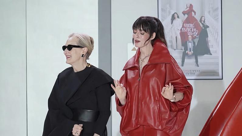 Duas pessoas em ambiente interno. À esquerda, indivíduo de roupa preta com cinto largo e óculos escuros; à direita, pessoa em traje vermelho de couro com zíper e acessórios dourados. Atrás deles, pôster com quatro figuras em trajes formais, incluindo uma em vestido vermelho sobre escada, com texto em coreano.