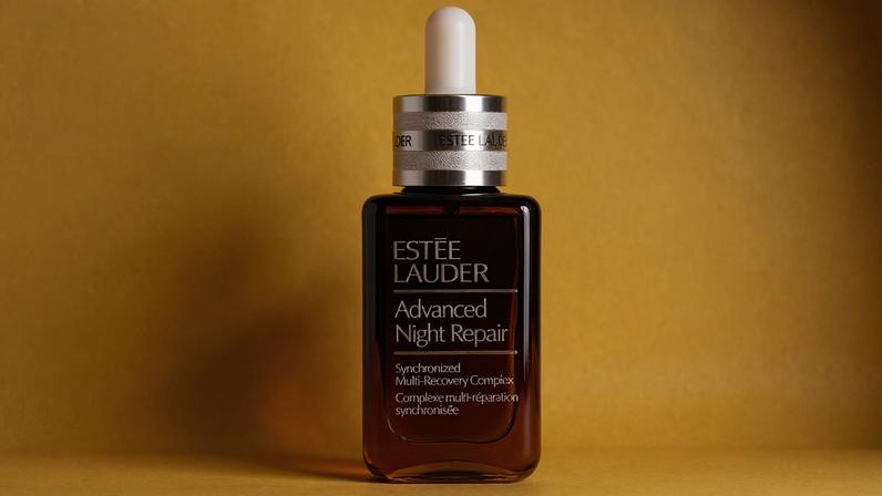 Estée Lauder advanced night repair serum bottle laying on a yellow background with yellow flowers surround it.