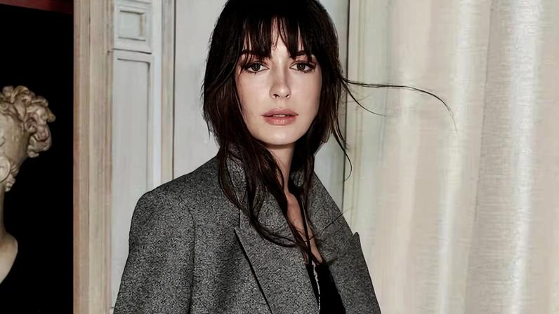 Anne Hathaway em foto colorida. Ela é uma mulher branca, de cabelo castanho. Usa uma blusa preta e um blazer cinza