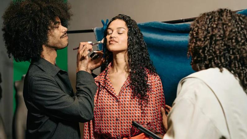 A person with long, dark curly hair and their eyes closed sits for a makeup application. A makeup artist with a large afro and a nose ring stands to the left, using a brush to apply powder to the person's cheek. To the right, another individual with curly hair, seen from behind, assists with the process. The person receiving makeup wears a red and black patterned shirt with a matching belt. The background includes a green screen and a blue fabric backdrop draped over a clothing rack.