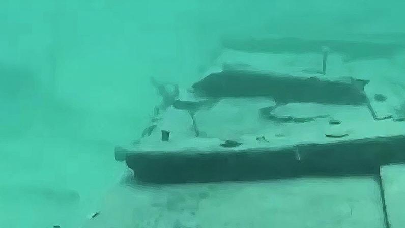Estrutura submersa coberta por água esverdeada, possivelmente um destroço ou objeto afundado, vista de cima no fundo do mar ou de um rio