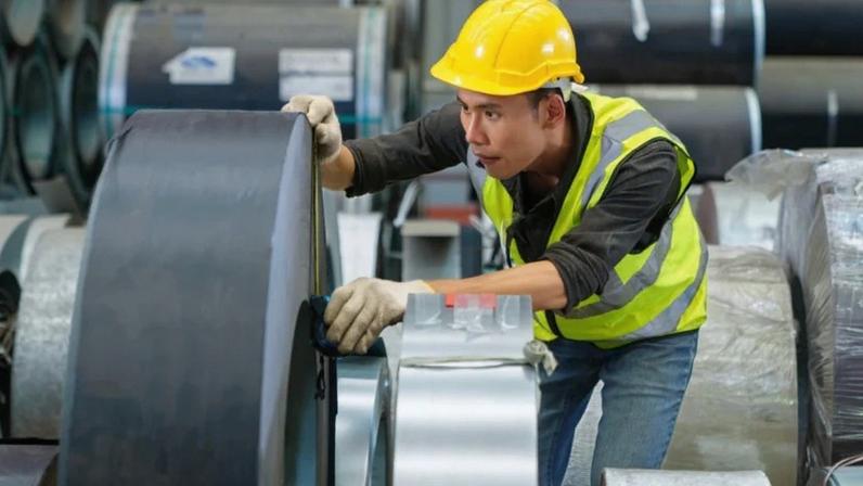 Foto de banco de imagens de operário inspecionando bobina de chapa metálica em armazém industrial.