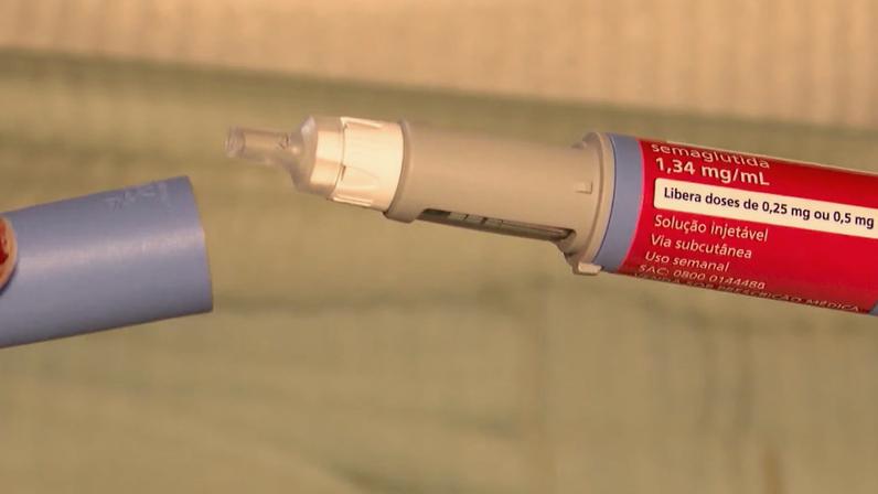 Caneta injetora médica com rótulo vermelho e tampa azul sendo retirada. Texto visível indica medicamento semaglutida, solução injetável de uso semanal por via subcutânea. Cena mostra detalhe de dispositivo utilizado para aplicação de doses controladas.