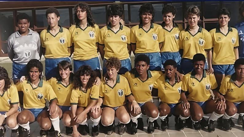 Foto de equipe de futebol em duas fileiras. Jogadoras vestem uniforme amarelo com gola verde, shorts azuis e meias brancas, lembrando seleção brasileira. Sapatos pretos completam o traje. No grupo, goleira aparece em uniforme cinza e outro atleta em azul.