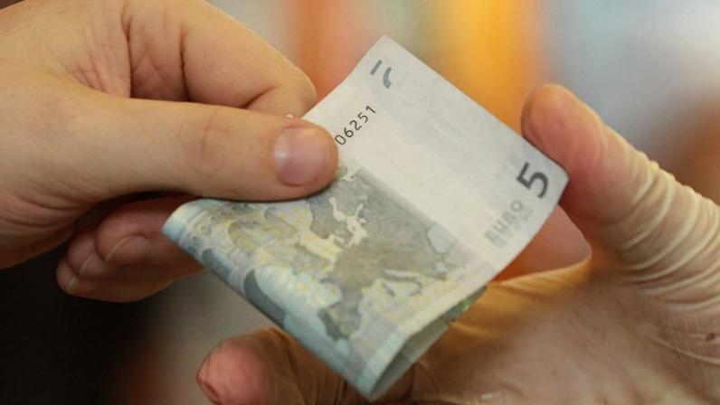 Uma mão sem luva entrega uma nota de 5 euros para uma mão enluvada.